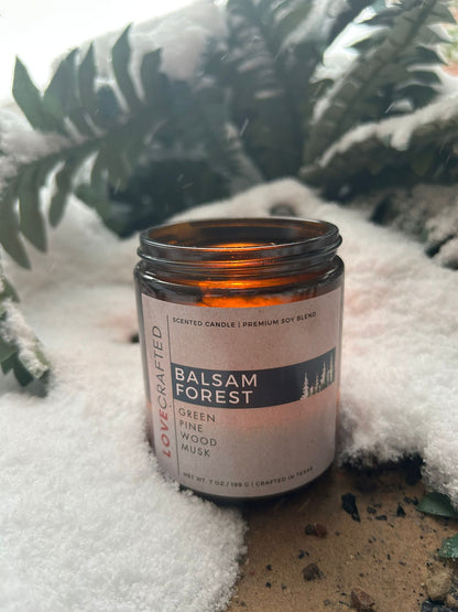 Lit Balsam Forest candle nestled in snow with evergreen branches