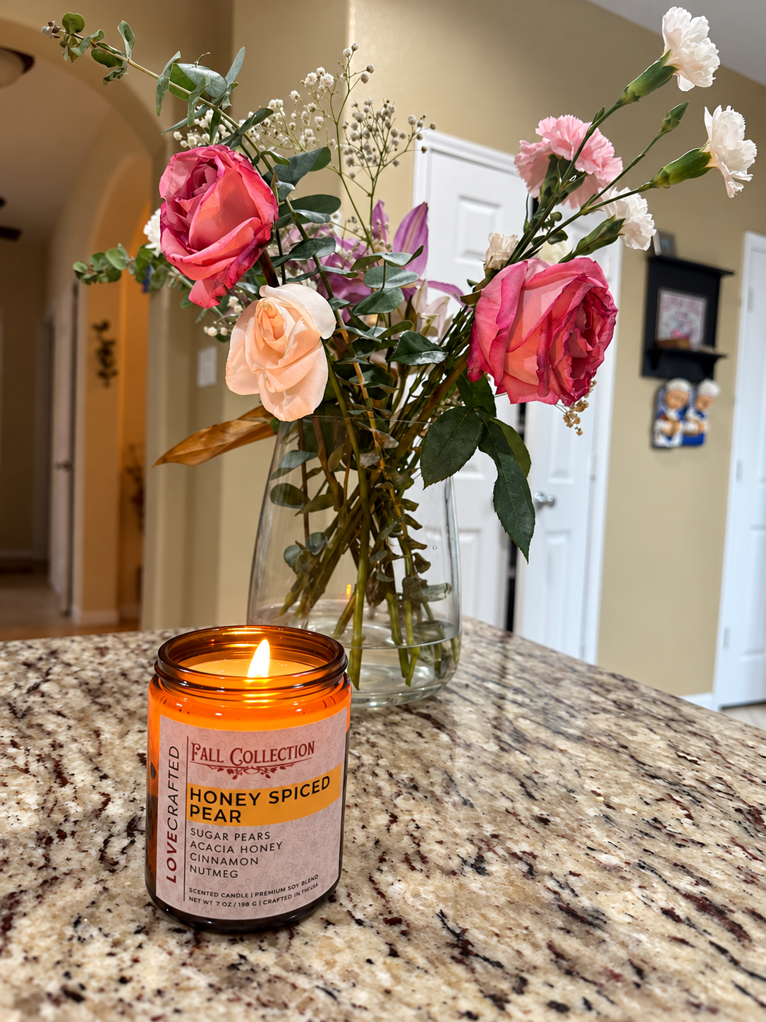 Lit Honey Spiced Pear candle on a kitchen counter beside a vase of pink roses