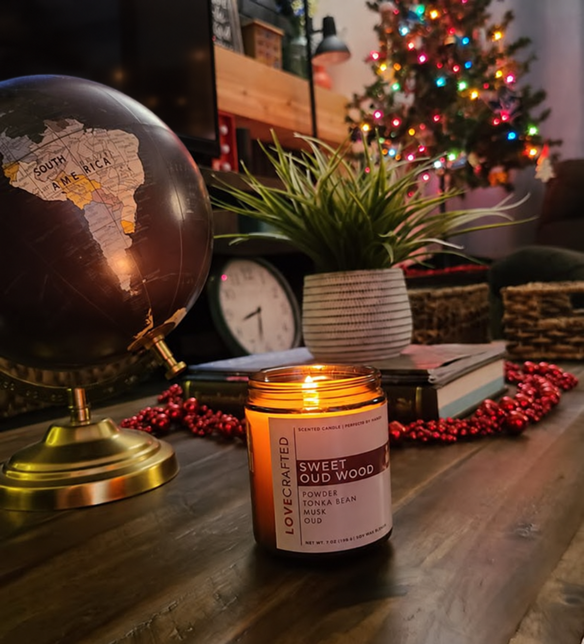 Lit Sweet Oud Wood candle on a desk with a globe and Christmas tree